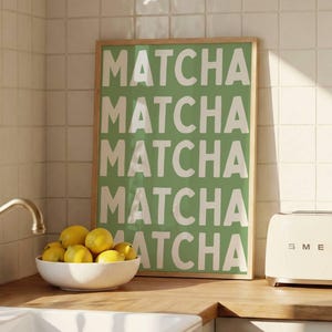 May include: A rectangular framed wall art print featuring the word MATCHA repeated five times in large white capital letters against a solid green background, displayed on a kitchen counter next to a bowl of lemons and a cream toaster.