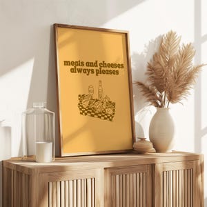 A framed golden yellow print featuring dark brown text that says meats and cheeses always pleases above a charcuterie picnic illustration.