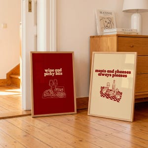 May include: Two rectangular framed wall art prints. The left print is deep red with white line art of wine glasses and snacks, featuring the text 'wine and picky bits'. The right print is cream with red line art of a picnic, reading 'meats and cheeses always pleases'.