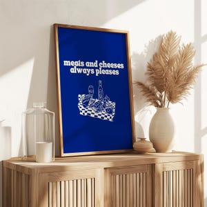 A framed royal blue print featuring white text that reads meats and cheeses always pleases above a charcuterie picnic illustration.