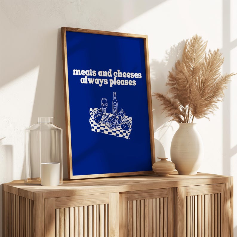 A framed royal blue print featuring white text that reads meats and cheeses always pleases above a charcuterie picnic illustration.