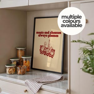 May include: A framed wall art print with a cream background and dark red line art illustration of a picnic basket containing bread, cheese, and wine. The text reads, "meats and cheeses always pleases" in a bold, dark red, rounded font.