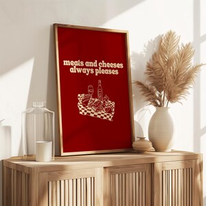A framed deep red print displaying white text that says meats and cheeses always pleases and a charcuterie picnic illustration.