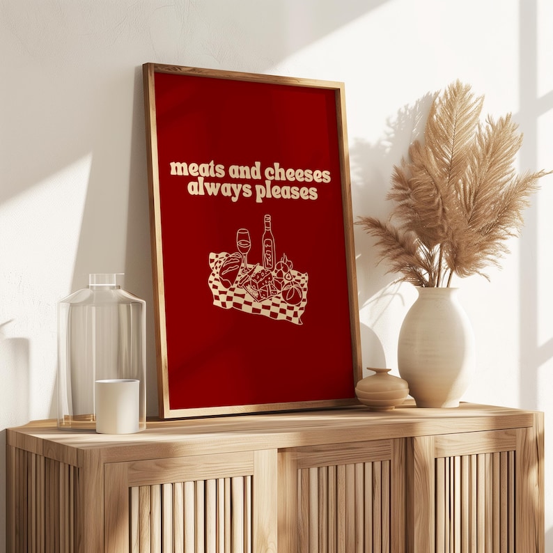 A framed deep red print displaying white text that says meats and cheeses always pleases and a charcuterie picnic illustration.