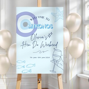 Puede incluir: Un letrero azul claro sobre un caballete, con el texto "Welcome to Mykonos" y "Olivia's Hen Do Weekend". El letrero incluye ilustraciones de una estrella de mar, peces y una copa de vino. Globos blancos en el fondo.