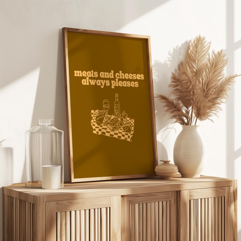 A framed mustard yellow print featuring white text that reads meats and cheeses always pleases above an illustration of a charcuterie picnic.