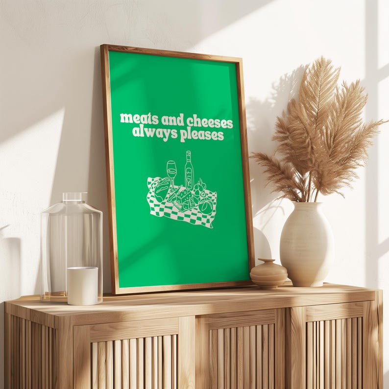 A framed vibrant green print showing white text that reads meats and cheeses always pleases alongside a charcuterie picnic graphic.