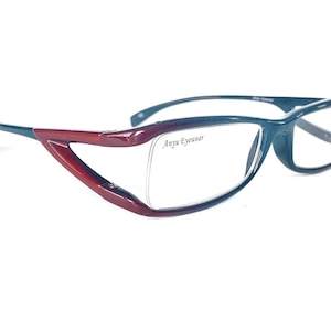 Op de afbeelding: Een paar donkergroene en bordeauxrode brillen met een heldere lens. Het montuur heeft een uniek ontwerp met een gebogen, hoekige vorm. De merknaam "Anya Eyewear" is op de rechterlens gedrukt.