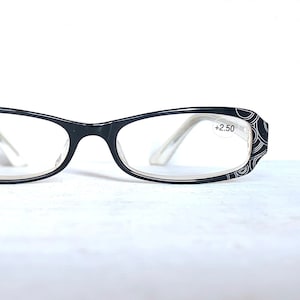 以下が含まれることがあります： 白いテンプルが付いた黒い長方形の眼鏡。フレームの外縁には装飾的な白い模様があります。右レンズには「+2.50」の文字があります。