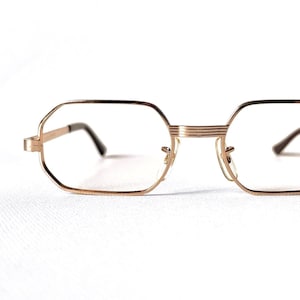Könnte beinhalten: Goldfarbene Brille mit achteckigem Rahmen. Die Brille hat klare Gläser und einen dünnen Metallrahmen. Die Bügel und der Steg sind ebenfalls goldfarben, was einen stimmigen und stilvollen Look erzeugt. Das Design ist von Vintage inspiriert.