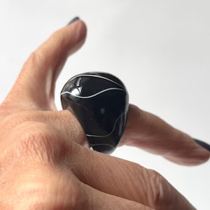 May include: A large, black ring with white, swirling patterns. The ring has a rounded, teardrop shape and is worn on a finger. A smaller, gold ring is visible on another finger. The background is plain white.