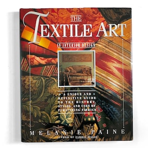 May include: A hardcover book titled "The Textile Art in Interior Design" with a colorful cover featuring fabric patterns and a room scene. The book is by Melanie Paine and includes text about furnishing fabrics.