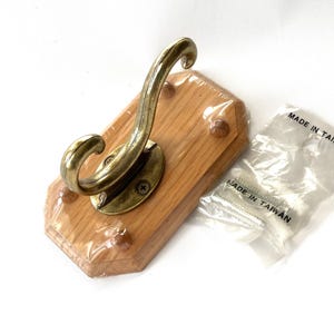 May include: A wooden wall hook with a brass-colored hook and mounting hardware. The hook is mounted on a light brown, rectangular wooden base. The packaging for the screws reads "MADE IN TAIWAN".