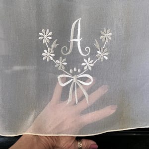 Peut inclure: Tissu blanc transparent avec des fleurs blanches brodées et une lettre "A" blanche au centre. Le tissu est plié en deux et tenu par une main.