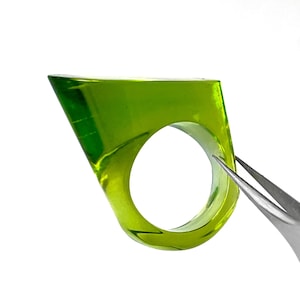 May include: A translucent lime green ring with a thick, angular, geometric silhouette. The ring features a smooth, polished surface and a circular opening for the finger, captured in a bright, clear light against a plain white background.