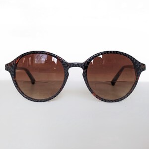 May include: Round bifocal glasses featuring a dark, textured frame with a subtle repeating pattern. The lenses are tinted in a gradient brown shade, and the bifocal line is clearly visible across the lower portion of each lens, providing a distinct visual feature.