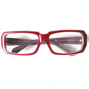 Op de afbeelding: Rode rechthoekige bril met de tekst "Anyu Eyewear" op de poot. De monturen zijn felrood en de glazen zijn helder. De bril heeft een retro, gedurfd ontwerp.