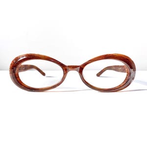 May include: Oval-shaped eyeglasses with a brown wood-grain pattern. The frames have a thick, rounded design, and the lenses are clear. The temples are also brown and feature the same wood-grain pattern. The glasses are set against a white background.