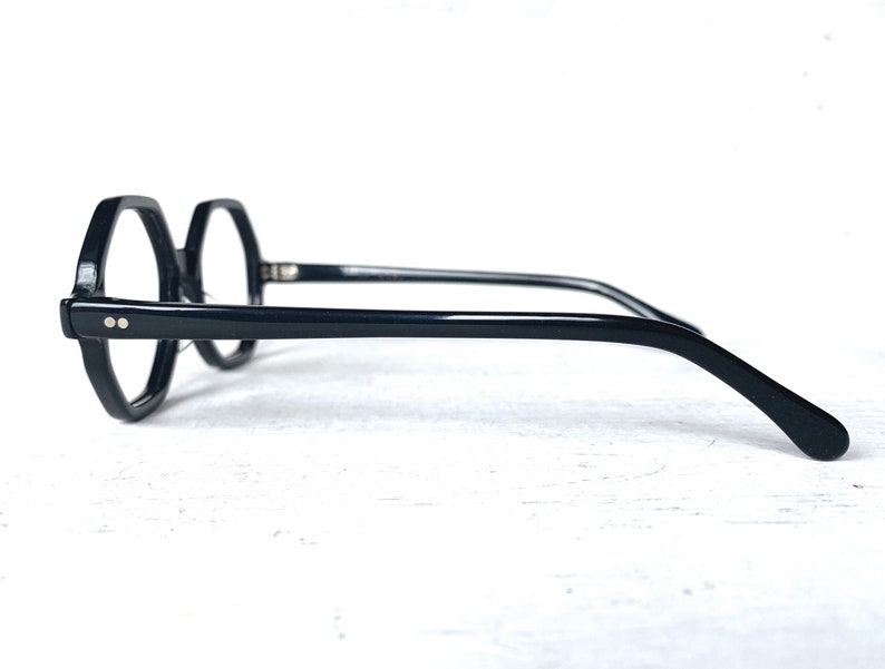 Puede incluir: Gafas negras con un dise&ntilde;o de montura octogonal &uacute;nico. Las gafas tienen un acabado brillante y presentan dos peque&ntilde;os remaches plateados en la parte delantera de la montura. Las patillas son elegantes y se curvan suavemente.