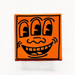 May include: Orange square artwork featuring a smiling face with three eyes and a black outline. The artwork has a minimalist design with bold lines and a cheerful expression. The background is white.