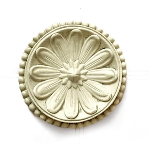 May include: A cream-colored, circular decorative element with a daisy-like design. The center features a raised, star-shaped detail, surrounded by large, curved petals. The outer edge is adorned with small, rounded beads, creating a textured border.