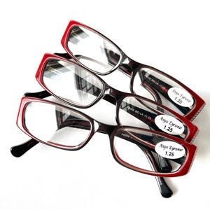 May include: Three pairs of rectangular reading glasses with red frames and black temples are stacked on a white surface. Each pair has a label that reads "Anyu Eyewear 1.25". The glasses have clear lenses.