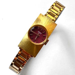 May include: A vintage gold-toned Tressa watch with a rectangular face and a burgundy dial. The watch features a gold-toned band and a textured, brushed finish on the watch face. The watch is a classic timepiece.