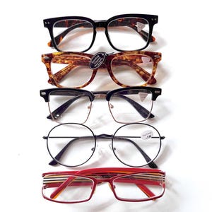 May include: Five pairs of spectacles are arranged on a white surface. The frames are in various styles and colours, including black, tortoiseshell, and red. Some have round frames, while others are rectangular or square.