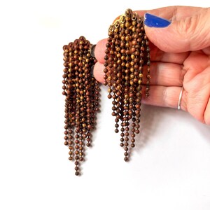 May include: A pair of vintage clip-on earrings with a cascade of small, brown beads. The earrings have a gold-toned metal backing and are held against a white background. The beads hang in a fringe-like pattern.