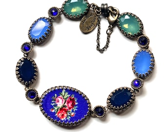 Puede incluir: Una pulsera con cristales ovalados facetados en tonos azules y verdes engarzados en bronce antiguo. La pieza central es una placa ovalada grande con fondo azul y un estampado floral de rosas rosas y rojas. Una pequeña etiqueta dice Michal Negrin.