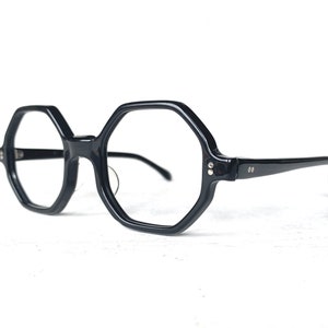 Puede incluir: Gafas negras de forma geom&eacute;trica con montura gruesa. Las lentes son transparentes y las patillas son finas. Las gafas tienen un estilo retro y vintage.