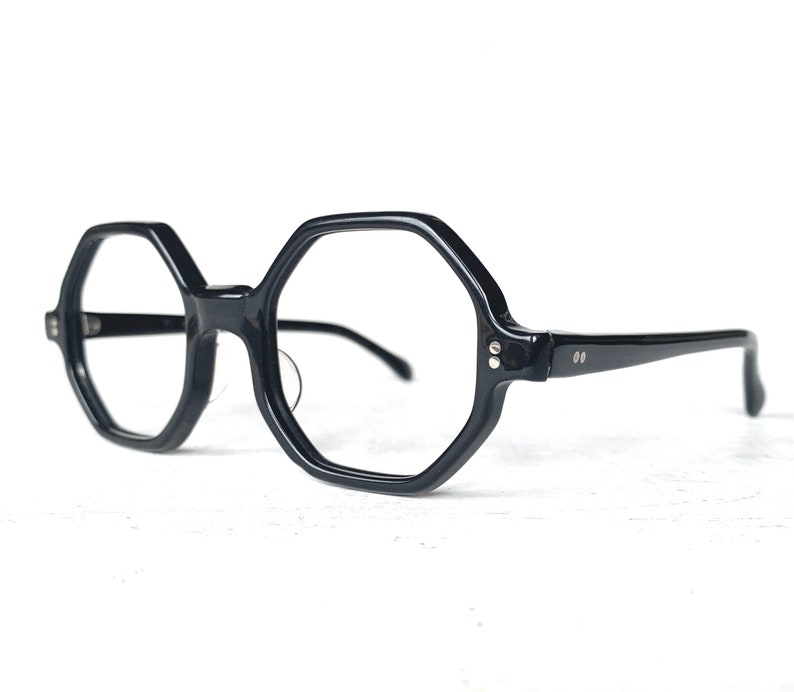 Puede incluir: Gafas negras de forma geom&eacute;trica con montura gruesa. Las lentes son transparentes y las patillas son finas. Las gafas tienen un estilo retro y vintage.