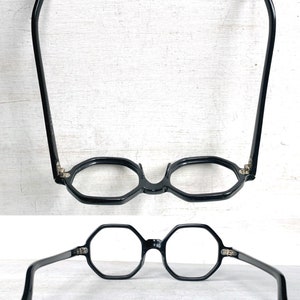 Puede incluir: Gafas negras octogonales con puente de cerradura y patillas curvas. Las gafas se muestran desde dos &aacute;ngulos diferentes sobre un fondo blanco. Las monturas son de color negro brillante y las lentes son transparentes.