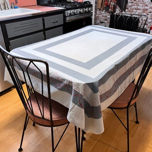 May include: A white tablecloth with a gray and red striped pattern and a gray border. The tablecloth is on a black metal table with two black metal chairs.
