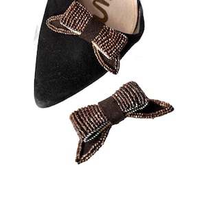 May include: A black pointed-toe shoe featuring a brown beaded bow accent on the vamp, accompanied by a separate matching brown beaded bow accessory. The shoe has a light-colored interior lining with the partial brand name Sam visible in gold lettering.