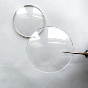 May include: Two clear, circular glass lenses are displayed against a white background. One lens is positioned above the other, with a pair of tweezers partially visible, suggesting a close-up view of the lenses.