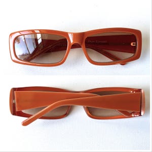 May include: Orange rectangular sunglasses with brown tinted lenses. The sunglasses have a thick frame and the text "Anyu Eyewear" is visible on the right lens. The arms of the sunglasses are also orange.