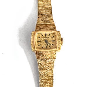 May include: A gold-toned wristwatch with a textured band and rectangular face. The watch face displays black hour markers and hands, with the brand name "WITTNAUER" visible. The watch band has a textured, bark-like appearance.