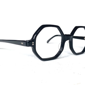 Puede incluir: Gafas negras de forma geom&eacute;trica con montura gruesa. Las gafas tienen un dise&ntilde;o octogonal &uacute;nico y est&aacute;n sobre un fondo blanco. Las patillas son finas y se extienden hacia los lados.