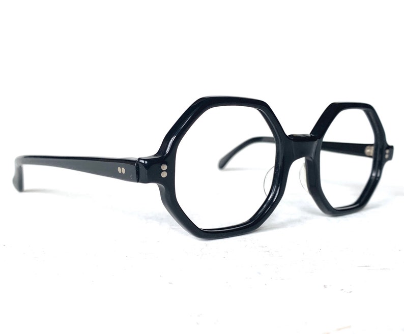 Puede incluir: Gafas negras de forma geom&eacute;trica con montura gruesa. Las gafas tienen un dise&ntilde;o octogonal &uacute;nico y est&aacute;n sobre un fondo blanco. Las patillas son finas y se extienden hacia los lados.