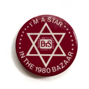 May include: A round, maroon button with white text and a white Star of David. The text reads "I'M A STAR" at the top, and "IN THE 1980 BAZAAR" at the bottom. The center has "BETH S BTS HOLOM" and "TEMPLE FLUSHING NY".