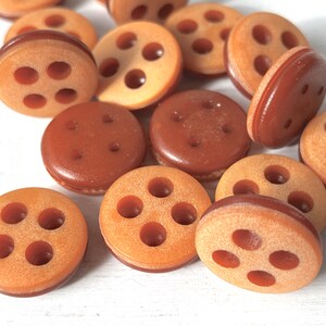 Könnte beinhalten: Eine Sammlung brauner und beiger Knöpfe mit jeweils vier Löchern. Die Knöpfe sind aus einem glatten, leicht strukturierten Material gefertigt.