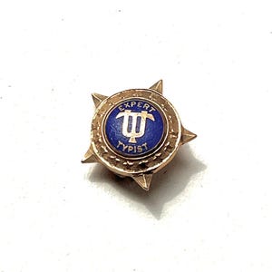 May include: A vintage gold-coloured pin with a blue centre. The centre has the words "EXPERT TYPIST" and a stylized letter "T". The pin has a starburst design around the edge.