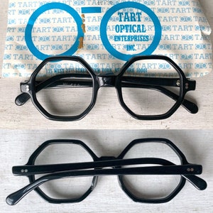 Puede incluir: Gafas negras con montura hexagonal y lentes transparentes, presentadas con su embalaje original. El embalaje presenta el texto "TART OPTICAL ENTERPRISES INC." y "HEXAGON BLACK". Las gafas se muestran tanto en posici&oacute;n vertical como invertida.