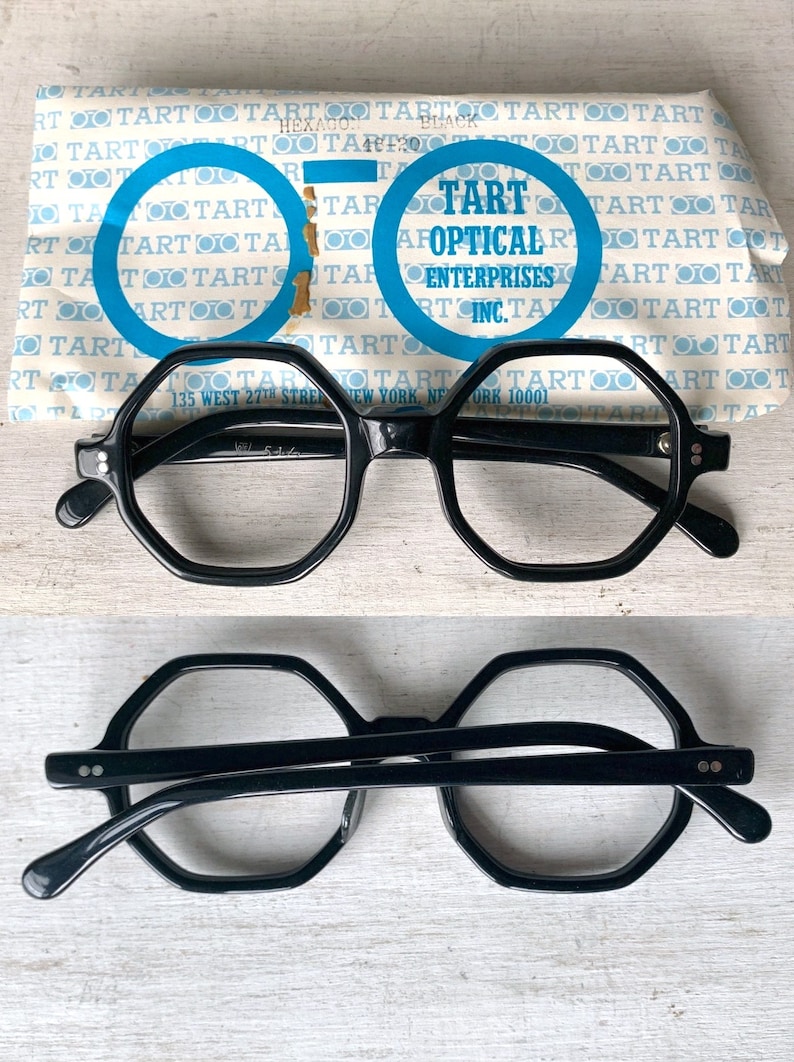 Puede incluir: Gafas negras con montura hexagonal y lentes transparentes, presentadas con su embalaje original. El embalaje presenta el texto "TART OPTICAL ENTERPRISES INC." y "HEXAGON BLACK". Las gafas se muestran tanto en posici&oacute;n vertical como invertida.