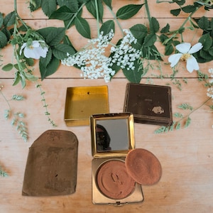 Può includere: Un compatto dorato con uno specchio e un batuffolo di polvere rotondo e marrone. Il compatto è aperto e il batuffolo è appoggiato sopra il compatto. Sullo sfondo ci sono tre scatole vuote, una con la scritta "Avon" stampata su di essa.