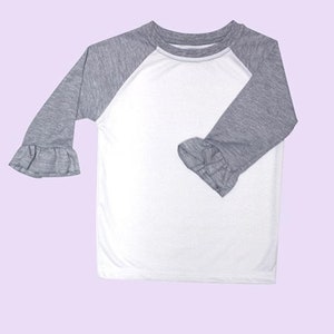 May include: A white and gray raglan style shirt with three-quarter length sleeves. The sleeves are gray with a ruffle detail at the wrist. The body of the shirt is white. The shirt is displayed on a light purple background.