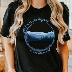 May include: Black t-shirt with a circular graphic featuring a mountain range and forest silhouette in shades of blue and purple. The text "Adventure begins where plans end" is arched around the design.