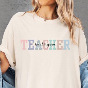 May include: Cream-colored t-shirt with the text "TEACHER third grade" in pastel colors. The word "TEACHER" is in pink, purple, and blue, while "third grade" is in black script. The shirt has rolled-up sleeves.
