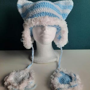 Può includere: Un cappello da gatto all'uncinetto a strisce blu e bianche con bordino in pelliccia bianca e guanti abbinati.